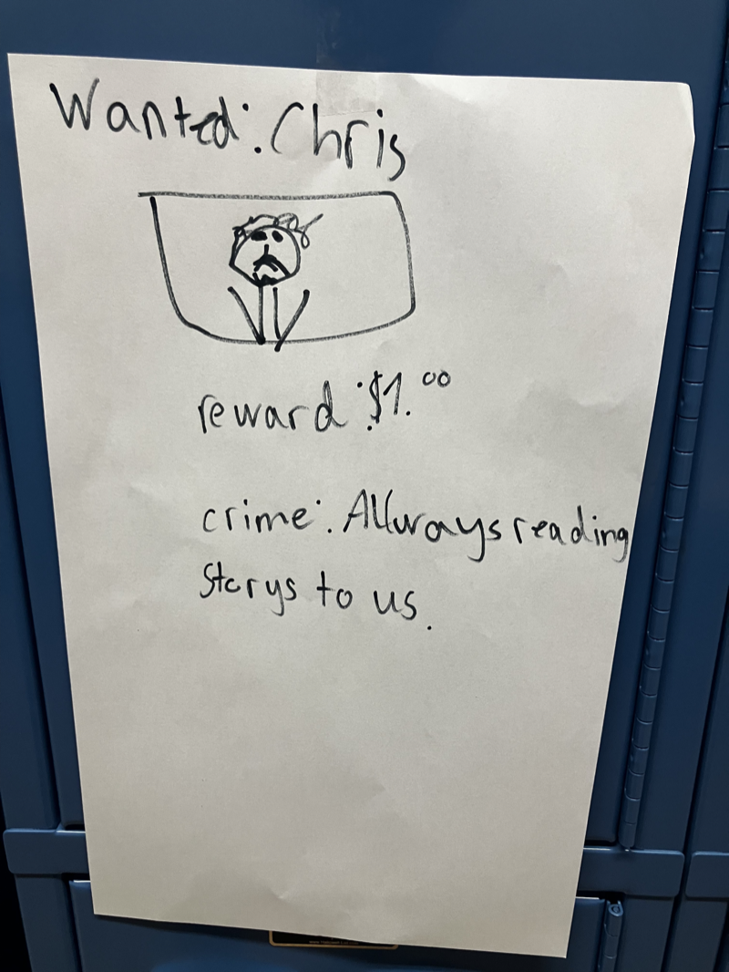 A student's hand-drawn note reading 'Wanted: Chris — reward $1.00 — crime: Always reading storys to us'